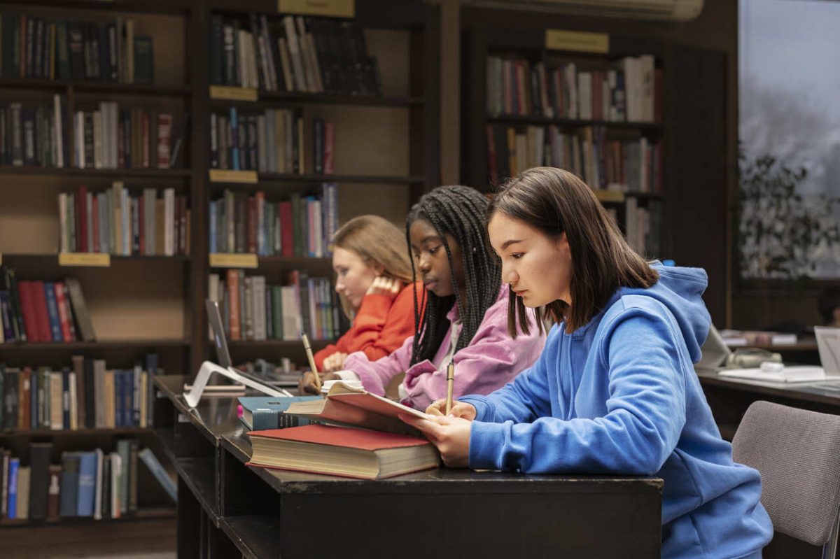 3 mulheres estudam juntas em biblioteca com cadernos, livros e canetas