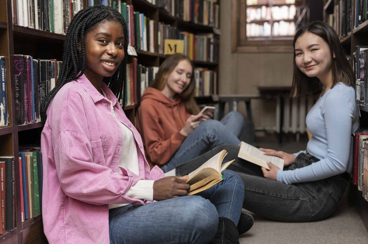 3 estudantes sentada no chão de biblioteca com livros 
