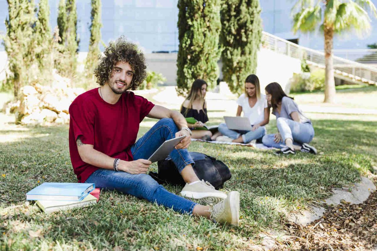 Estudante de camiseta vermelha sentado na grama com 3 estudantes com livros e cadernos