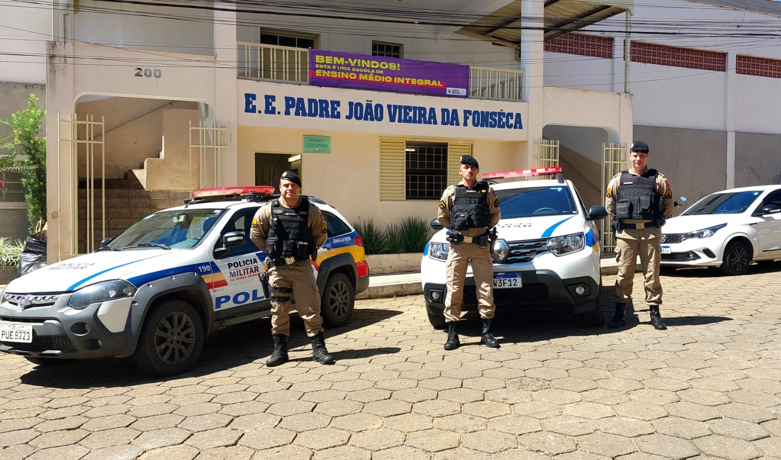 Três policiais militares de Minas Gerais à frente de suas viaturas, em frente uma escola estaduak