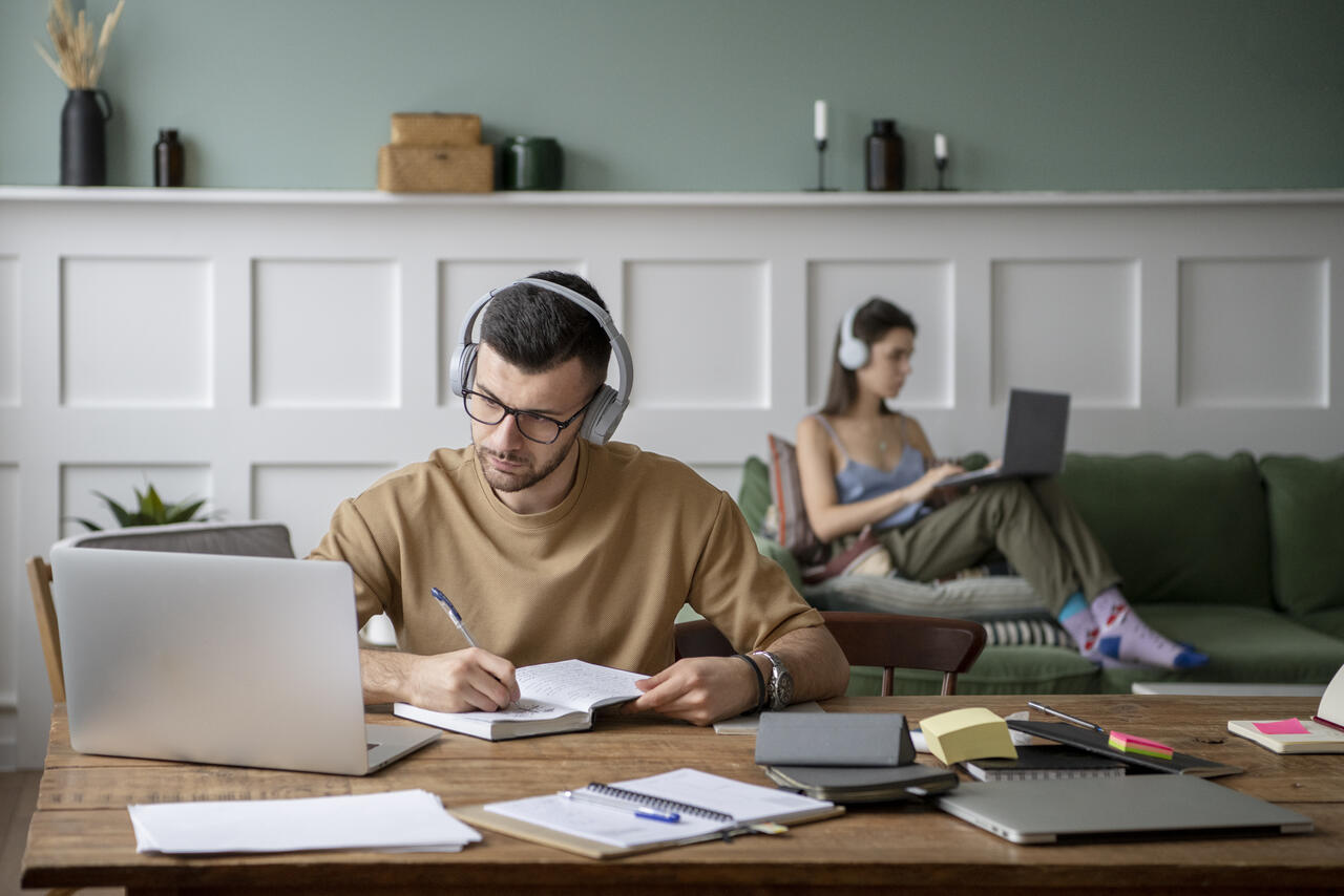 Homem e mulher estudam com computadores e fones de ouvidos em sala, com livros, anotações e canetas