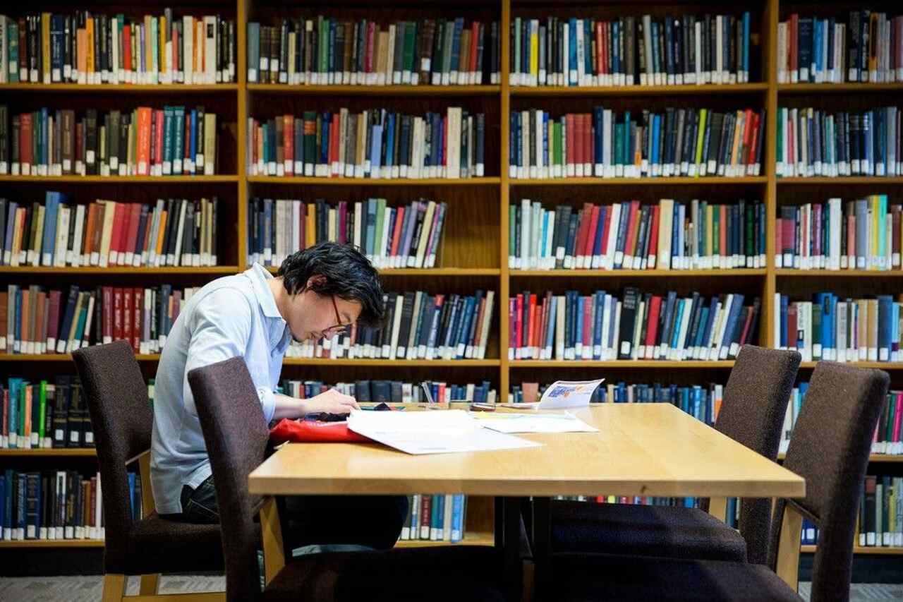 Aluno estudando com livros e cadernos sentado em mesa em biblioteca 