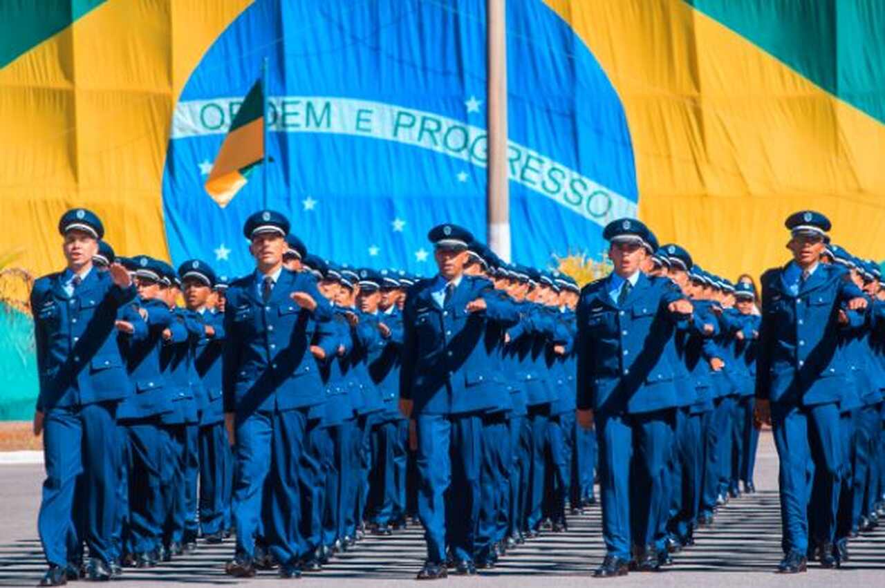 Sargentos FAB em fila de formação com bandeira do Brasil ao fundo