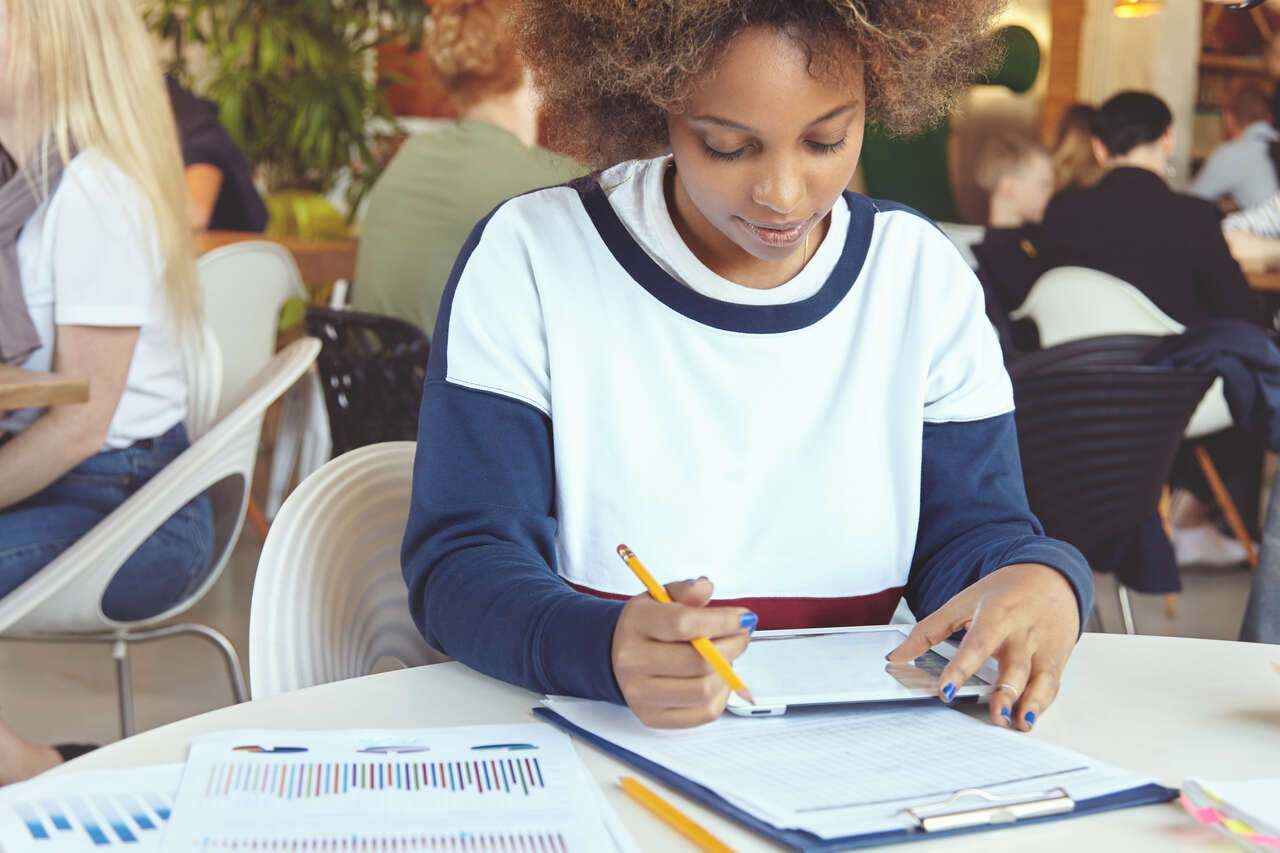 Jovem negra estudando sozinha em mesa de biblioteca com lápis e papéis 
